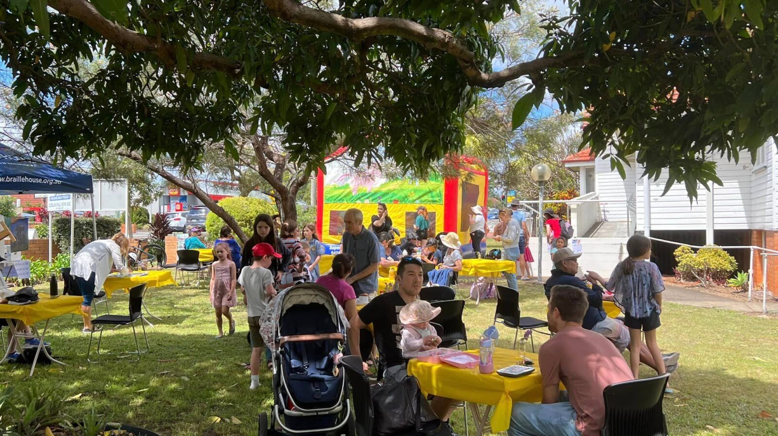 people sitting in the outdoors under a tree with a jumping castle in the background.