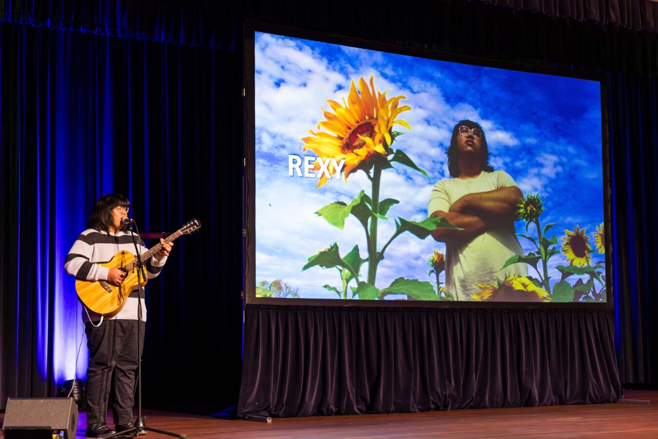 ng a guitar on a stage with a large screen behind them with a sunflower and a person